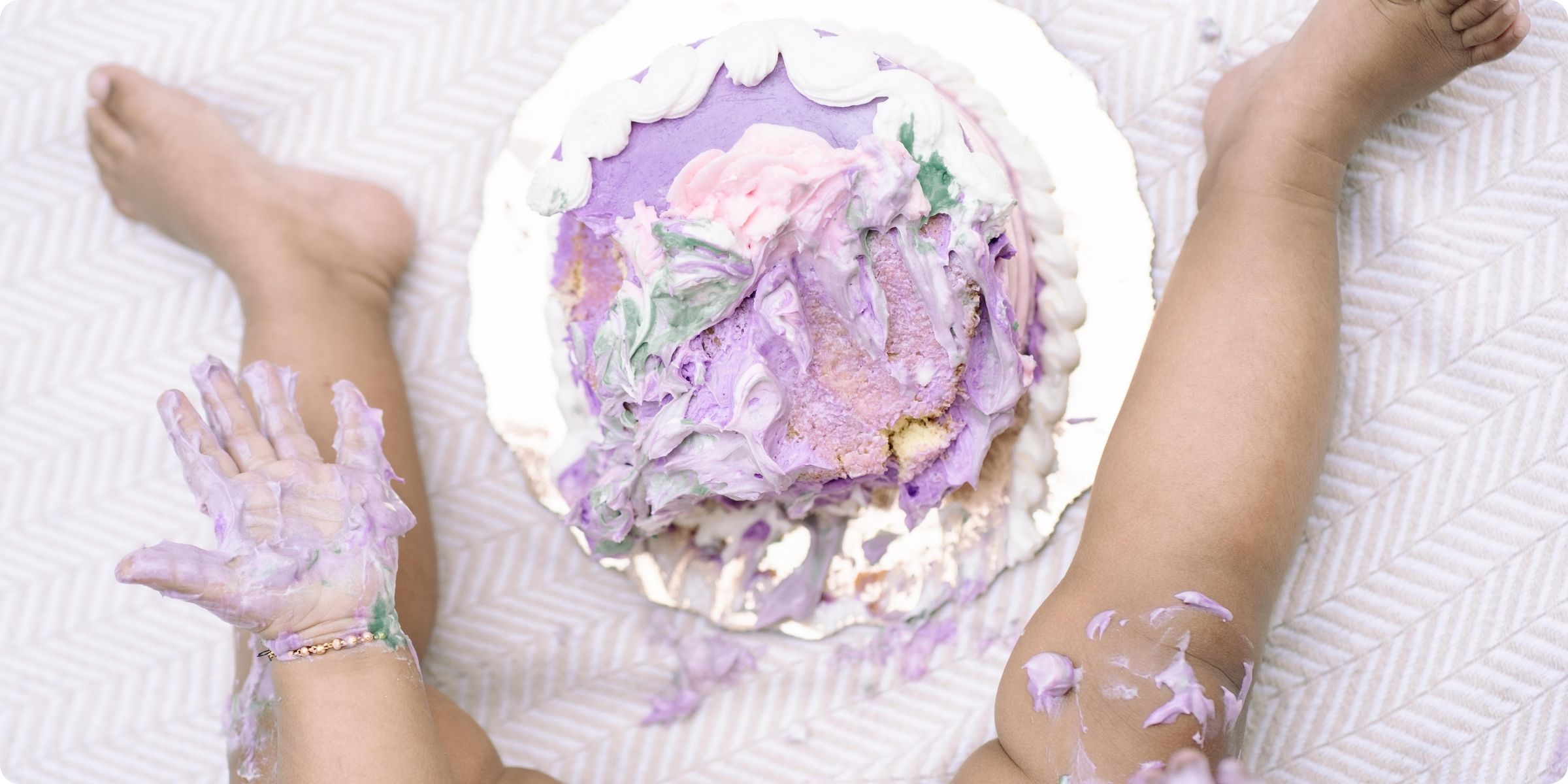 A top-down view of a first birthday cake smash in St. Pete, showing a toddler’s frosting-covered hands and legs alongside a partially smashed purple cake—capturing memories of playful, messy joy.