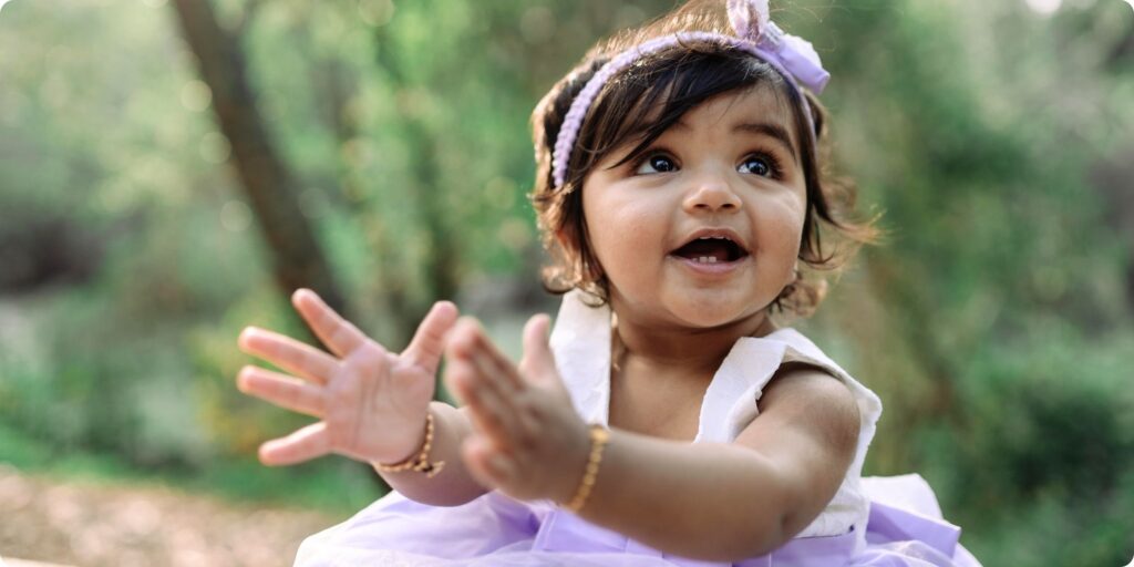 A baby girl in a lavender birthday dress claps her hands and beams with excitement during her first birthday photography session in St. Pete, capturing natural light, joy, and playful expression.