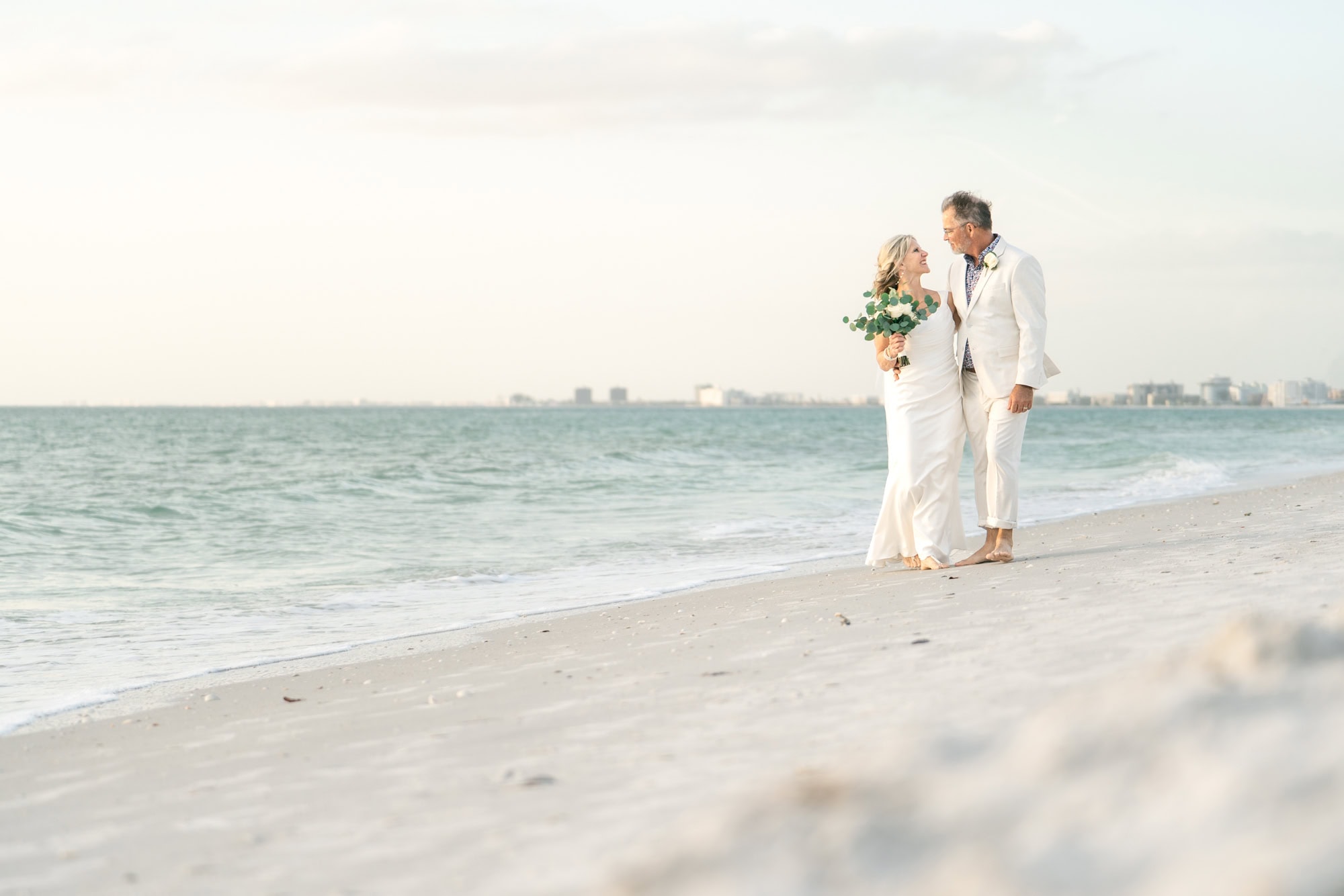 A bride and groom walk down the beach together as they head from their ceremony to their bell ringing