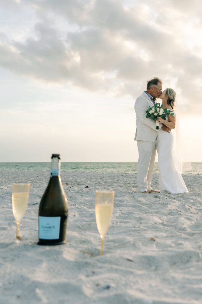 Champagne and glasses in the foreground of a newly married couple kissing on the beach