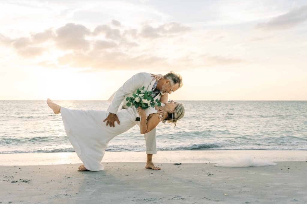 Just married couple dances and dips on the edge of the waves as the sun sets behind them