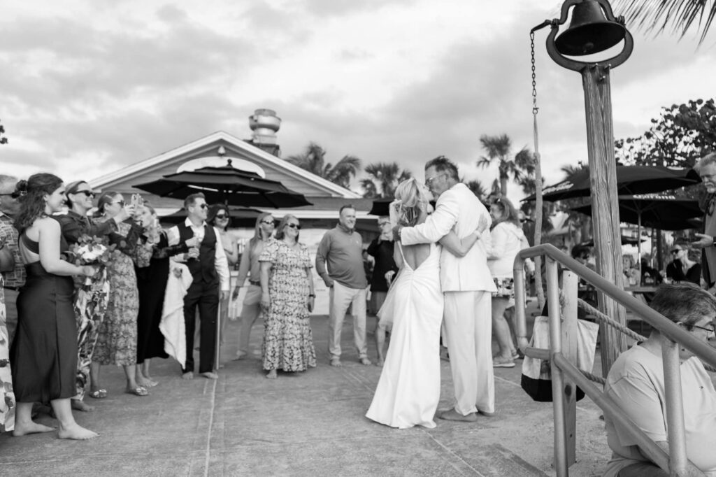 Newlyweds embrace surrounded by their friends and family on the patio right before ringing the bell at sunset