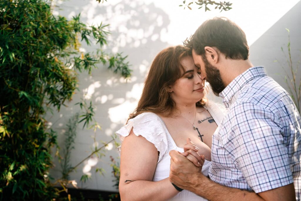 Tender close-up of a couple resting foreheads together in soft light with greenery around them. A natural engagement photo capturing emotion without stiff posing.