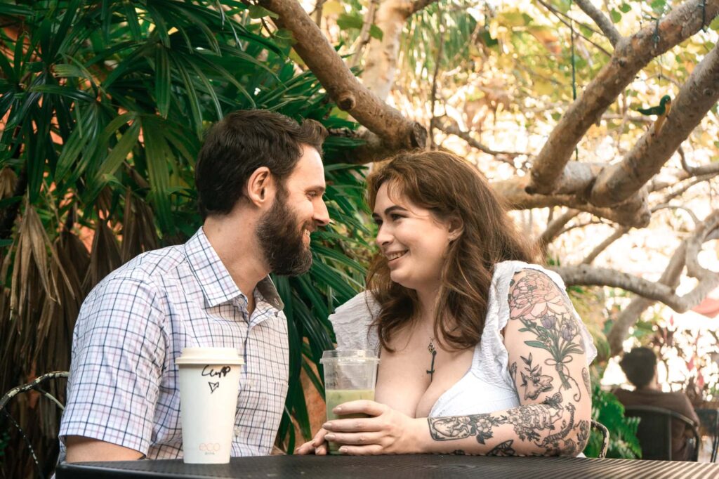 Smiling couple sitting at a café patio table with coffee cups, surrounded by greenery. A relaxed and candid St Pete engagement photo perfect for couples who feel awkward on camera.