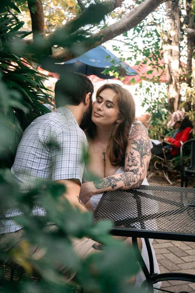 Couple leaning in for a kiss at a garden café table in St Petersburg, framed by plants. A playful and intimate engagement photo idea for couples who don’t love being photographed.