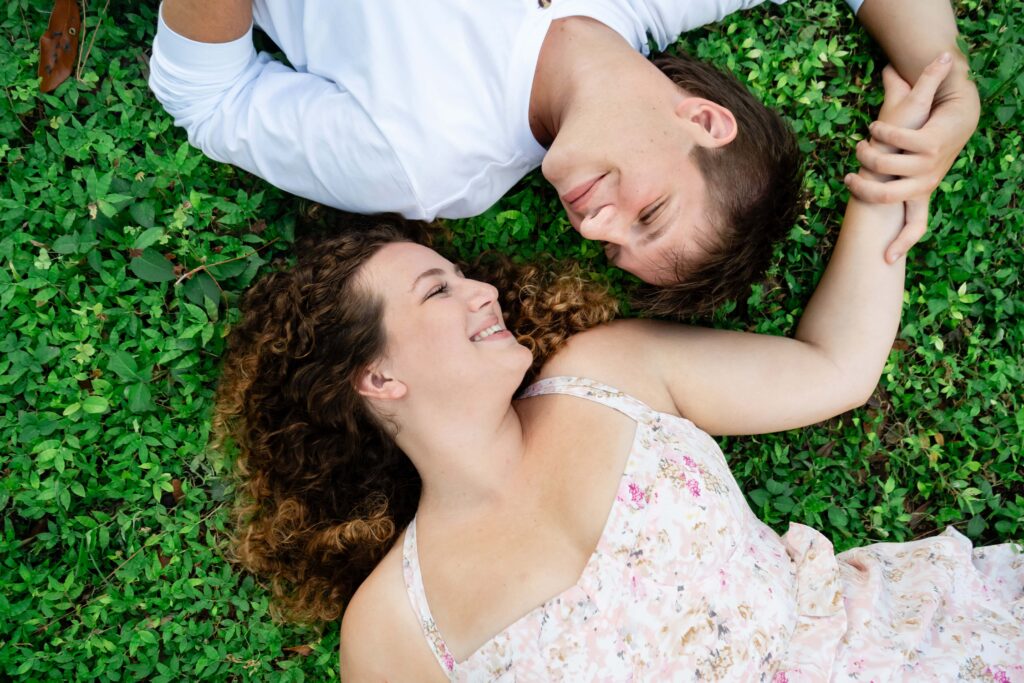 Couple lying in the grass holding hands and smiling at each other during their St Pete engagement session. A natural and candid moment showing how awkward engagement photos can turn fun and relaxed.