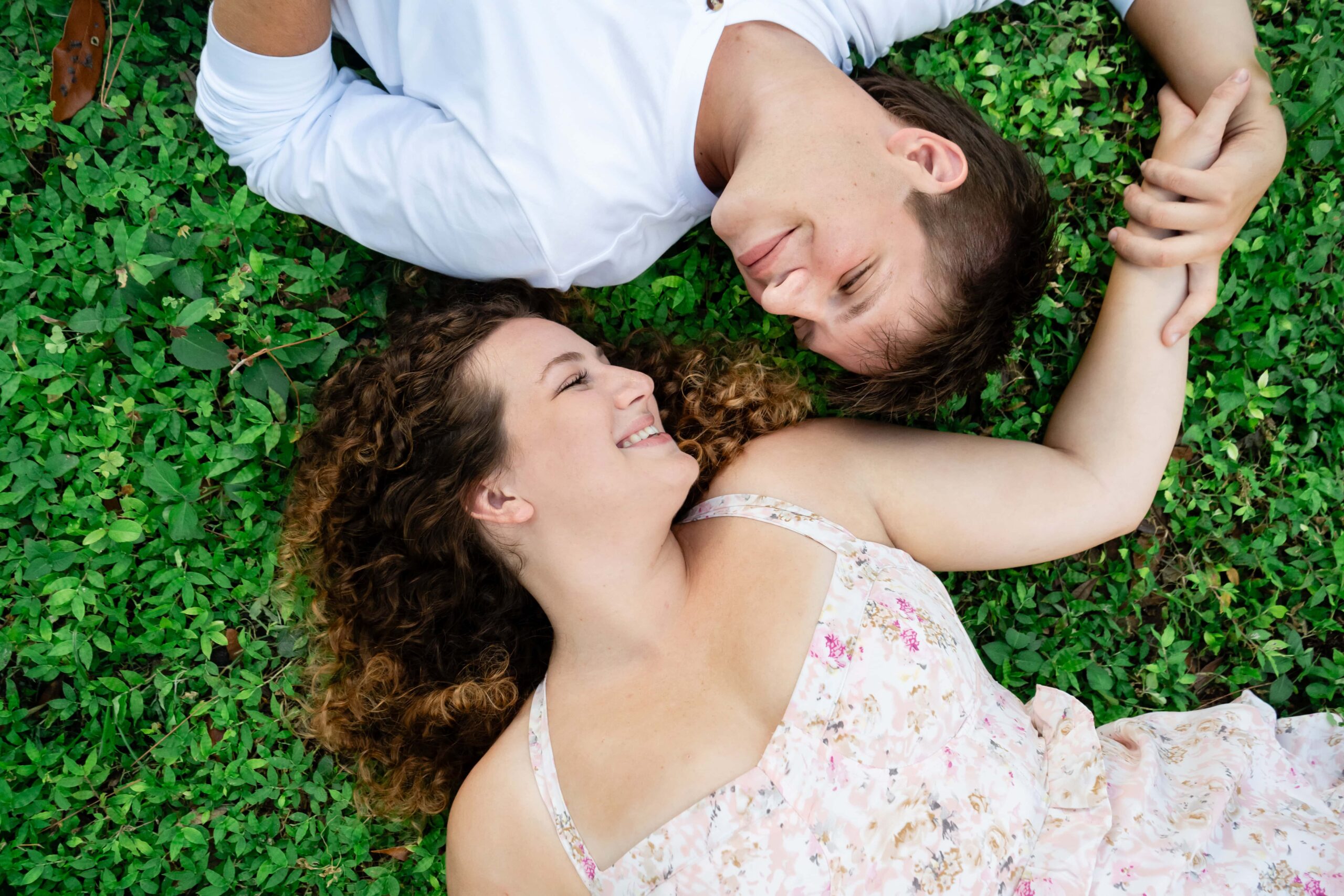 Couple lying in the grass holding hands and smiling at each other during their St Pete engagement session. A natural and candid moment showing how awkward engagement photos can turn fun and relaxed.