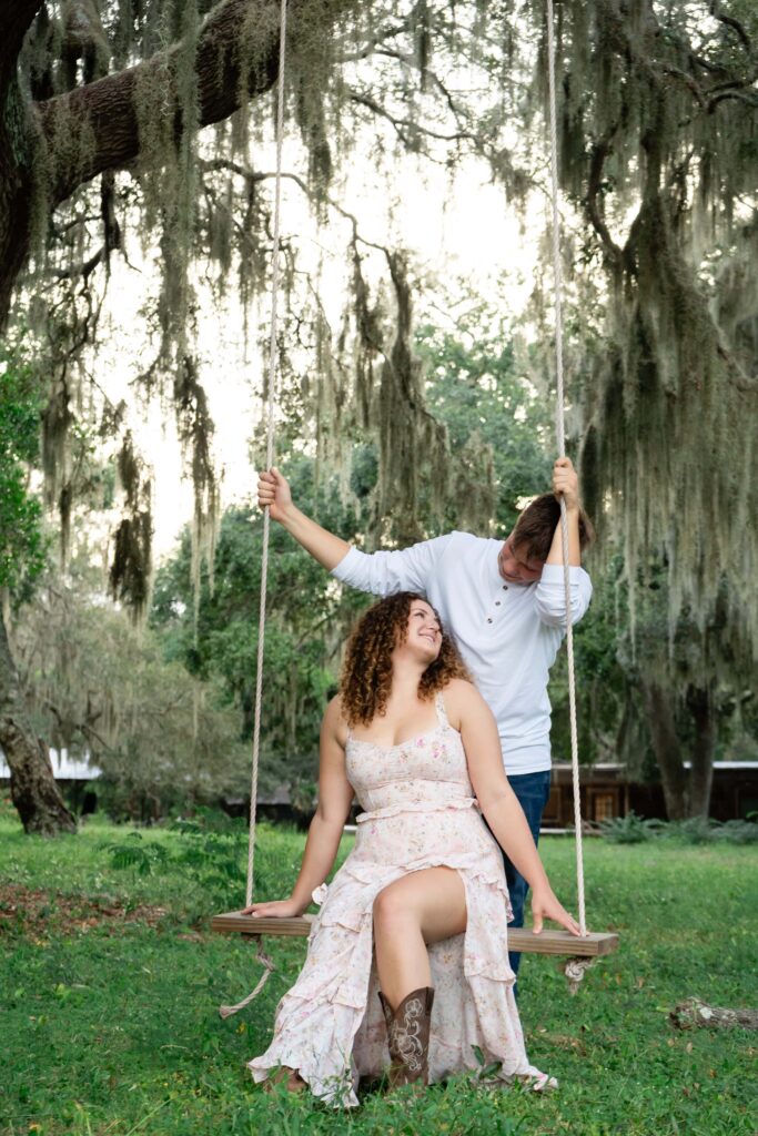 Engaged couple enjoying a playful moment on a wooden swing under Spanish moss in Florida. A relaxed, natural engagement photo in St Pete that feels fun instead of awkward.