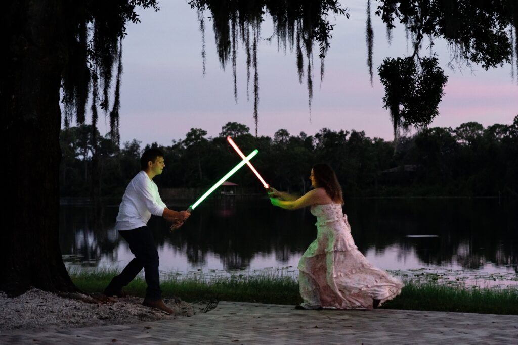 Playful nighttime engagement photo of a couple having a lightsaber duel by the lake in St Petersburg, FL. A unique and fun way to make awkward engagement photos feel personal.