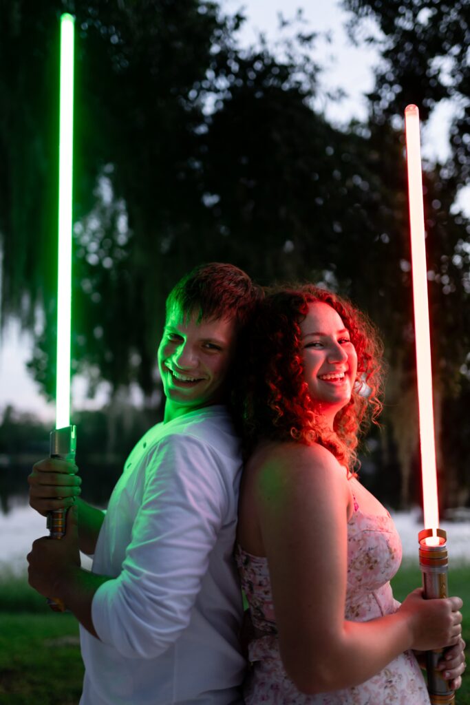 Engaged couple standing back-to-back, smiling while holding glowing lightsabers. A quirky and candid St Pete engagement photo idea for couples who don’t want traditional poses.