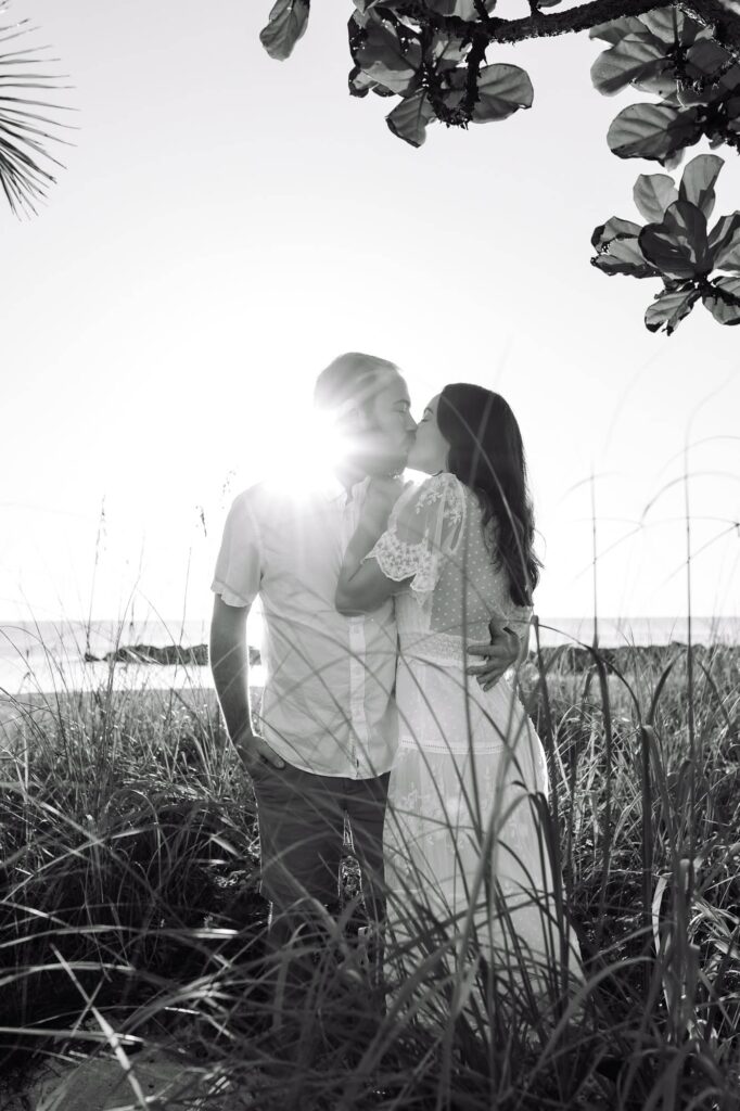 Black and white photo of a couple kissing on the beach in St Petersburg, FL, framed by sea oats and backlit by the sun. A romantic engagement photo idea for awkward couples who want candid, natural images.