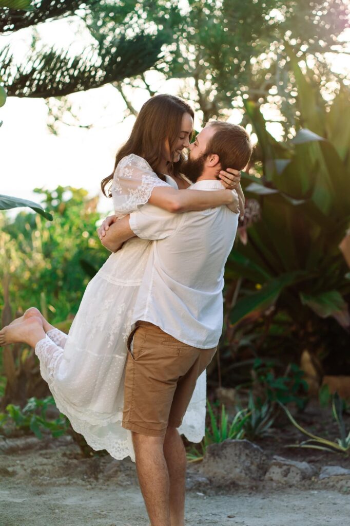 Man lifting his fiancée in the air while they kiss, surrounded by greenery. A joyful and playful St Pete engagement photo showing how awkward couples can look natural on camera.