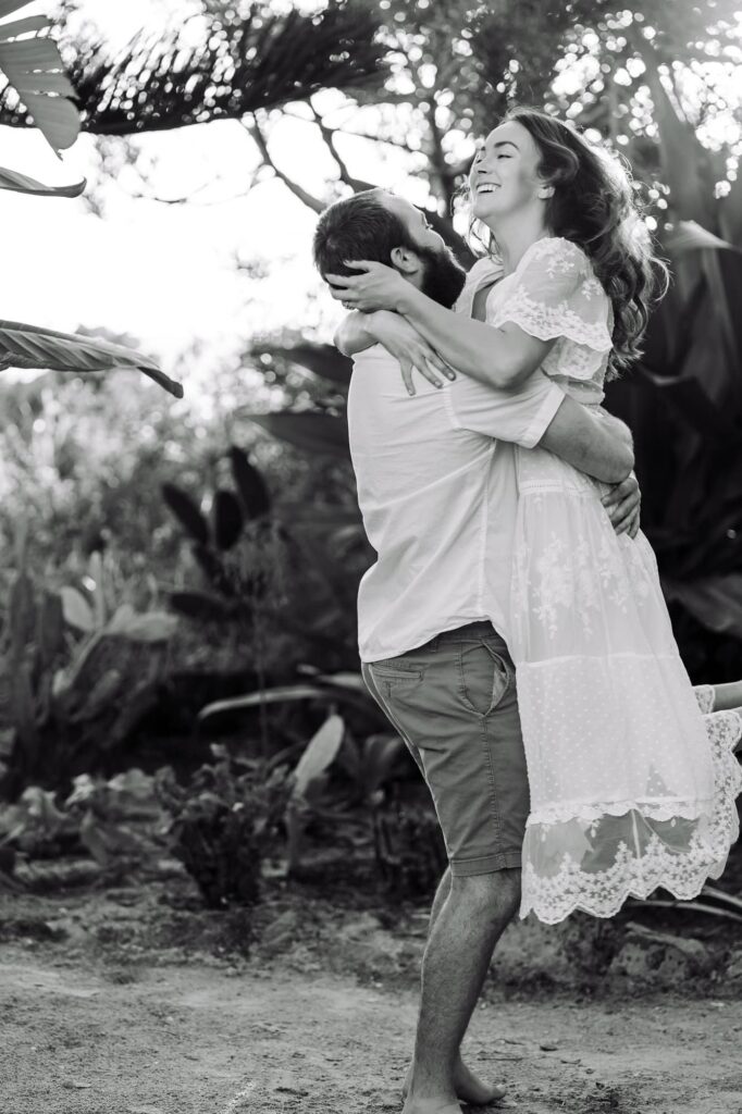 Black and white photo of a man lifting his fiancée and making her laugh under the trees. A candid engagement session idea in St Pete for couples who want relaxed, natural photos.