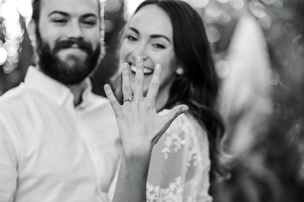 Woman holding out her hand to show off her diamond engagement ring, smiling at the camera while her partner looks on. A fun St Pete engagement photo idea that makes awkward posing feel natural.