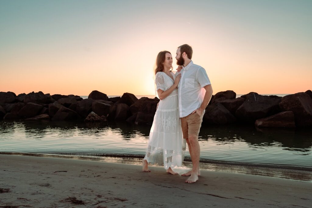 Couple standing barefoot on the beach at sunset near the rocks in St Petersburg, FL. A romantic engagement photo idea for couples who want natural, not awkward, portraits.