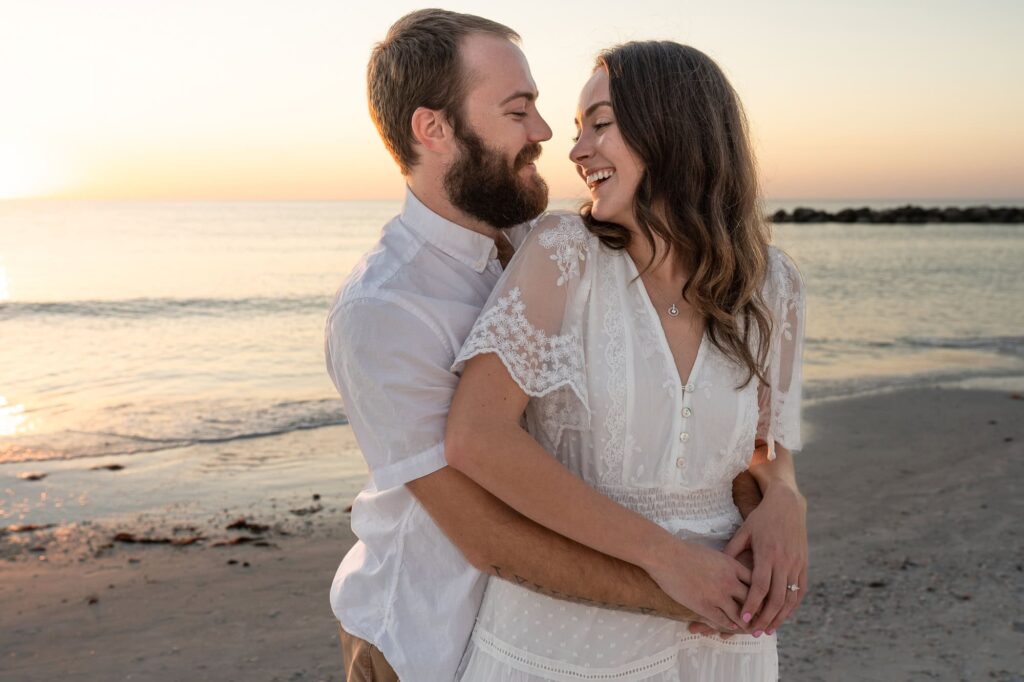 Man hugging his fiancée from behind as they laugh together during sunset on a St Pete beach. A candid engagement photo capturing fun, natural connection for couples who feel awkward posing.