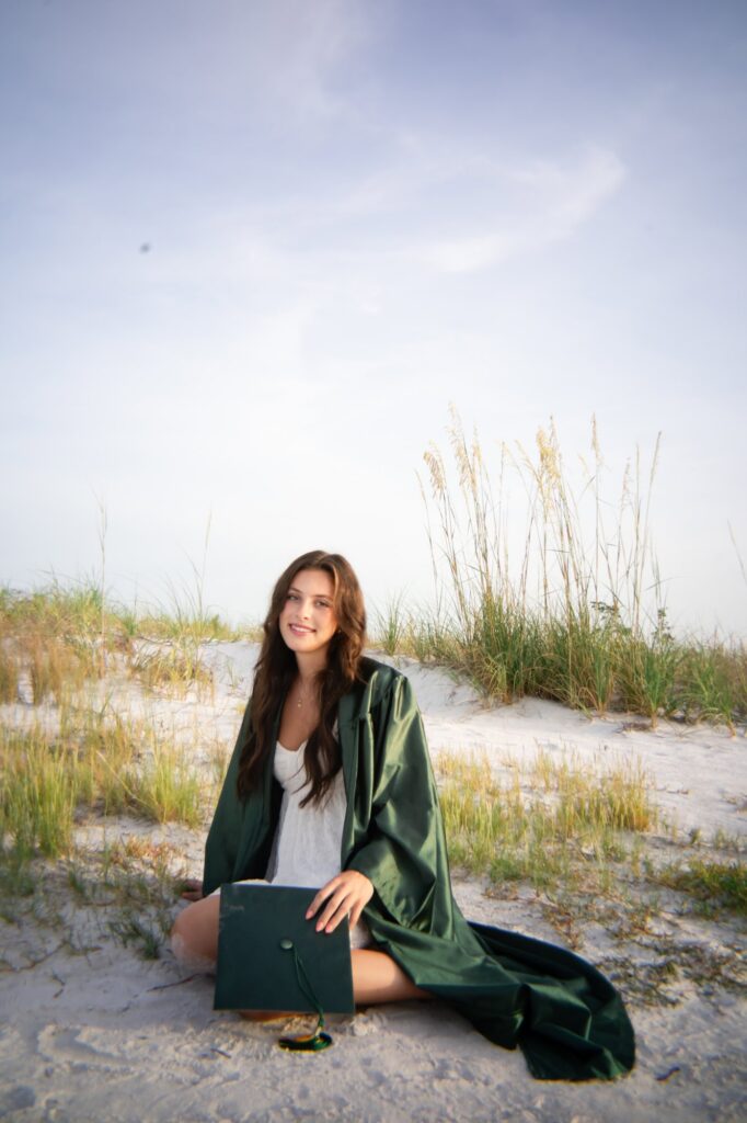 Graduating senior in green cap and gown sitting among beach dunes at Fort De Soto