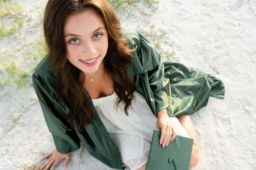 Senior girl in cap and gown smiling up toward the camera on the beach