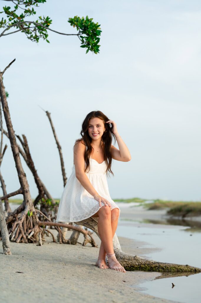 Teen sitting barefoot on driftwood in a white dress during senior portraits at the beach
