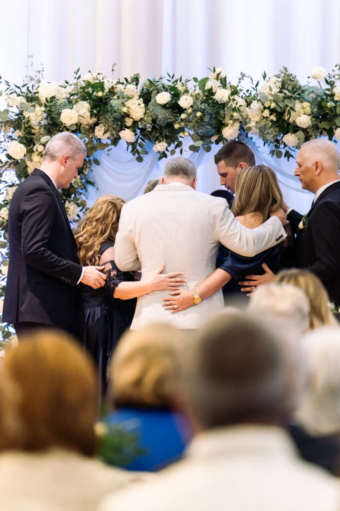 Family and couple sharing emotional prayer during wedding ceremony under floral arch, photographed by St. Pete area wedding photographer Erin Vestal.