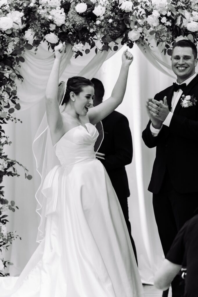 Bride celebrating joyfully with raised arms at end of wedding ceremony while groom applauds beside her, photographed by Erin Vestal Photography.