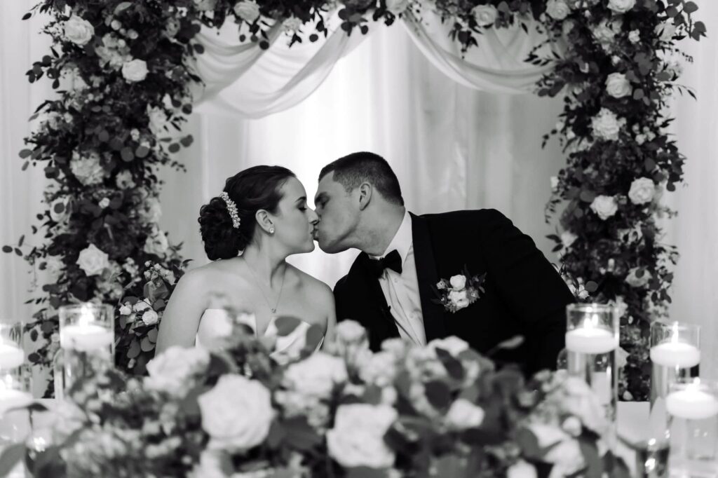 Bride and groom sharing a kiss behind floral sweetheart table under romantic flower arch during wedding reception, photographed by Erin Vestal.