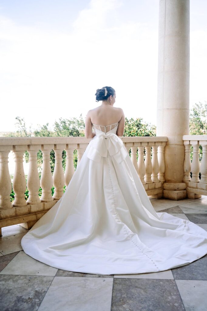 Bride standing on stone balcony in elegant satin gown with dramatic train, photographed by St. Pete wedding photographer Erin Vestal.