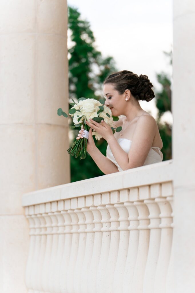 Bride smelling her bouquet of white roses and eucalyptus while standing on marble balcony, photographed by St. Pete wedding photographer Erin Vestal.