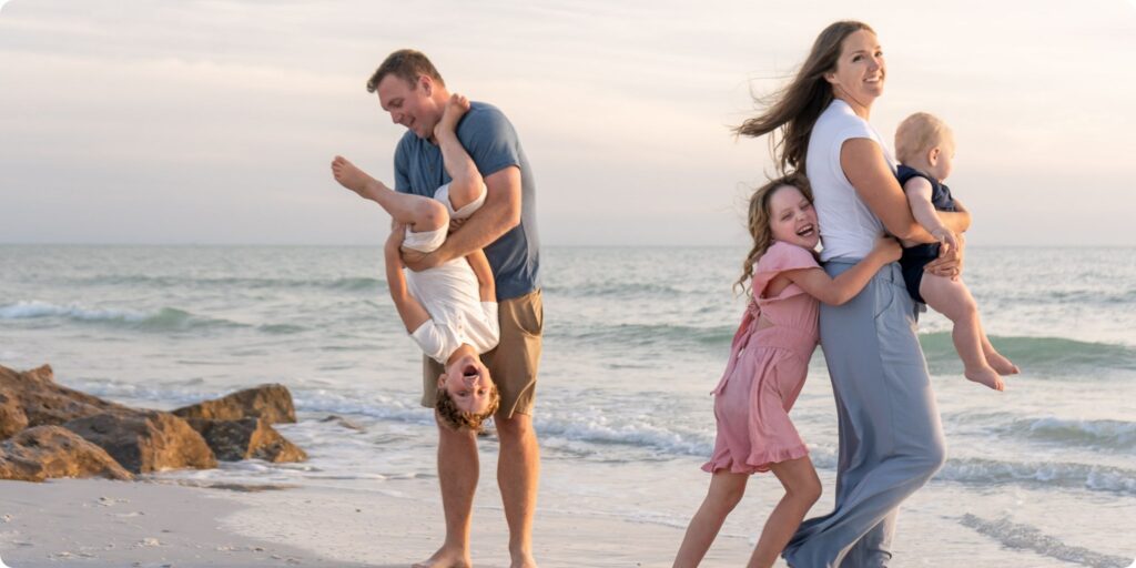 Family of five laughing together at Upham Beach, parents holding the baby while older kids hug and play in the surf.