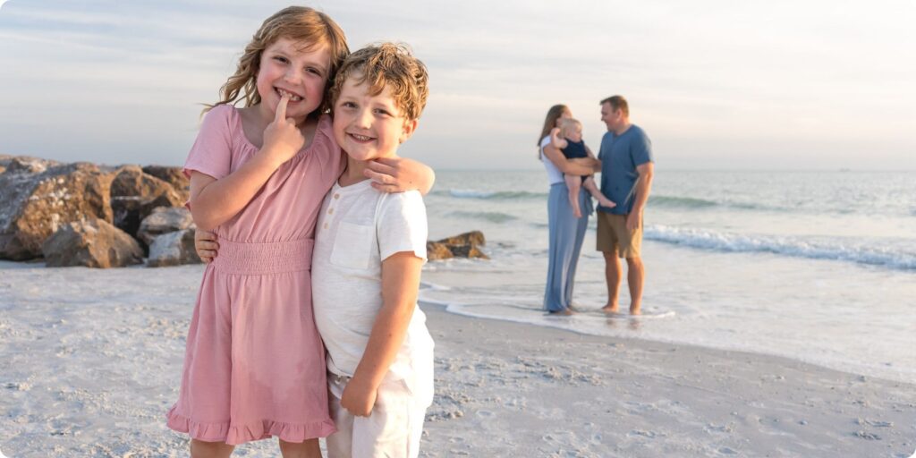 Siblings hugging and smiling at the camera with parents blurred in the background during candid family photos at Upham Beach.