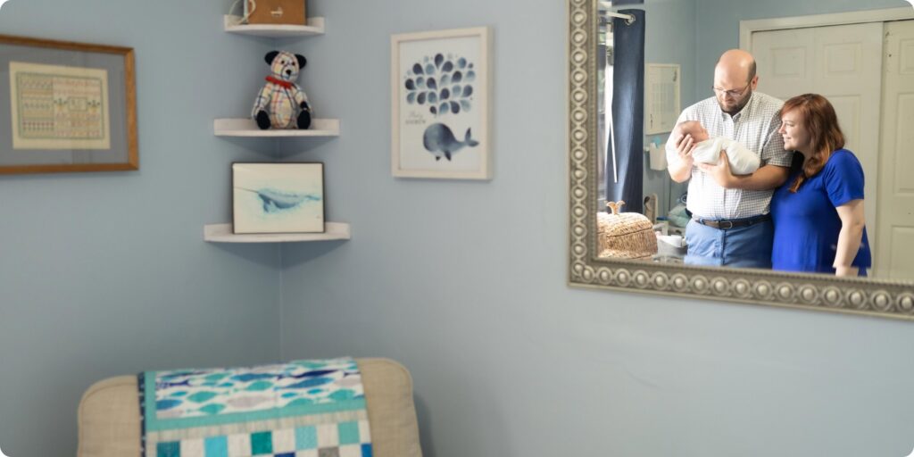 New parents reflected in nursery mirror holding their newborn, handmade quilt and childhood stuffed animals in the foreground — in-home newborn photography St. Petersburg