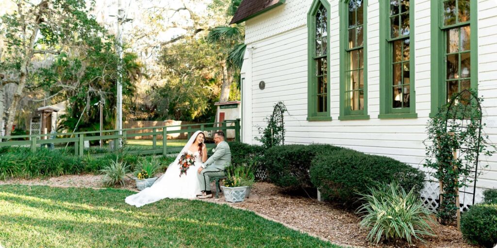 Bride and groom sitting together in the garden beside Historic Andrews Memorial Chapel in Dunedin Florida surrounded by palm trees and Spanish moss