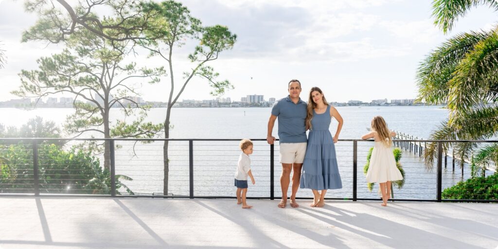 Family of four on a waterfront deck in Clearwater, Florida — parents standing together at the railing while toddler boy explores nearby and young daughter gazes out at the water