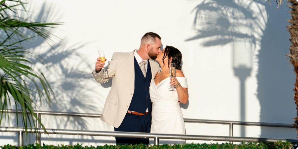 Bride and groom kissing on balcony at Opal Sands Resort Clearwater Beach holding champagne glasses with palm tree shadows