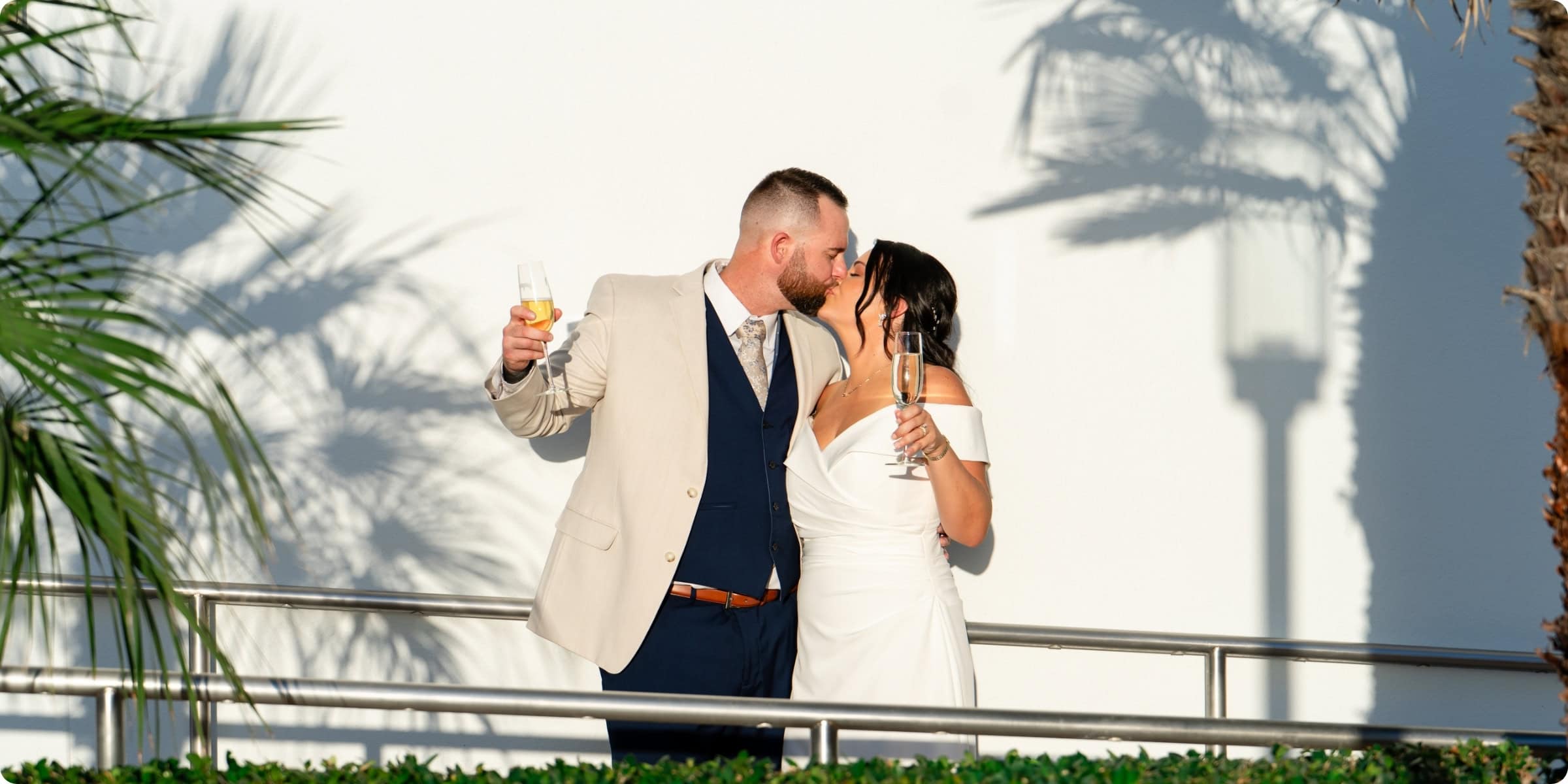 Bride and groom kissing on balcony at Opal Sands Resort Clearwater Beach holding champagne glasses with palm tree shadows