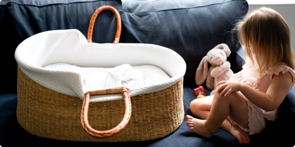 Toddler sitting beside a Moses basket with her stuffed bunny, taking her time warming up to her new baby sister during a St. Petersburg newborn session